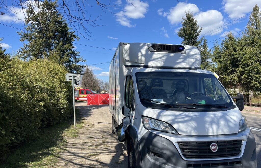 Biały samochód dostawczy zaparkowany przy drodze, z widoczną czerwoną bramą i wozem strażackim w tle.