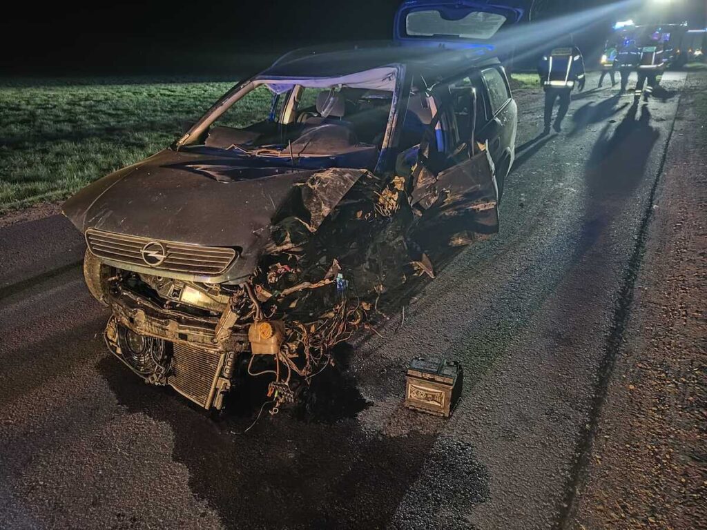 Zniszczony samochód osobowy stoi na drodze, obok widać interwencję służb ratunkowych.