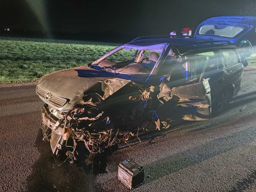 Uszkodzony samochód osobowy z wgniecionym przodem stoi na drodze, w tle widać oświetlenie służb ratunkowych.