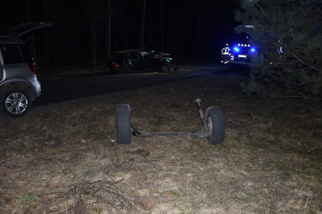 Uszkodzony samochód zaparkowany przy drodze, obok leży oderwana oś z kołami, w tle widać pojazdy służb ratunkowych.