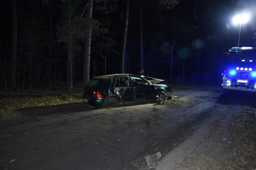 Uszkodzony zielony samochód osobowy stoi na leśnej drodze w nocy, obok niego widoczny wóz strażacki z włączonymi sygnałami.