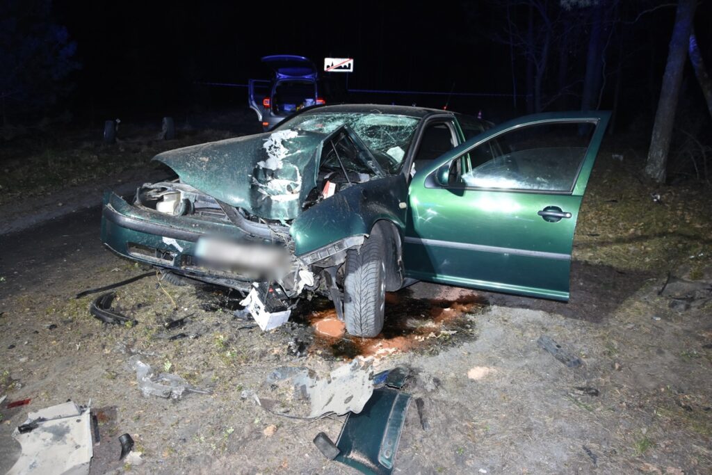 Zniszczony zielony samochód osobowy leży na poboczu drogi, z wgnieceniami na przedniej części i plamą oleju na ziemi.