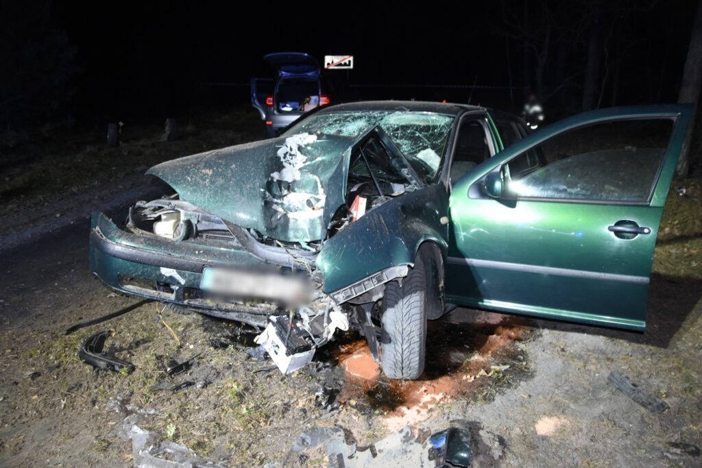 Zniszczony samochód osobowy leży na poboczu drogi, z wybitą szybą i uszkodzoną karoserią, w otoczeniu ciemności.