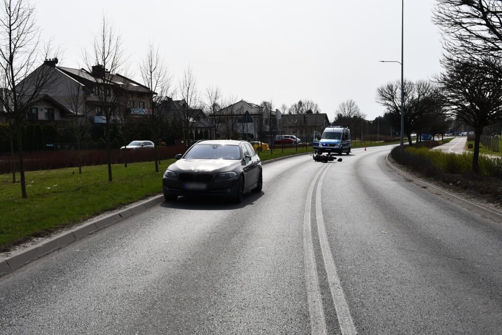 Czarny samochód osobowy zatrzymany na poboczu drogi, obok niebieski pojazd służbowy. W tle widać domy i drzewa.