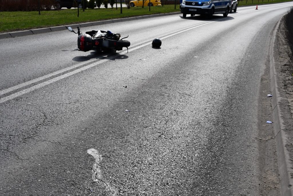 Motocykl leży na jezdni, obok kask, a w tle przejeżdżający radiowóz policyjny.