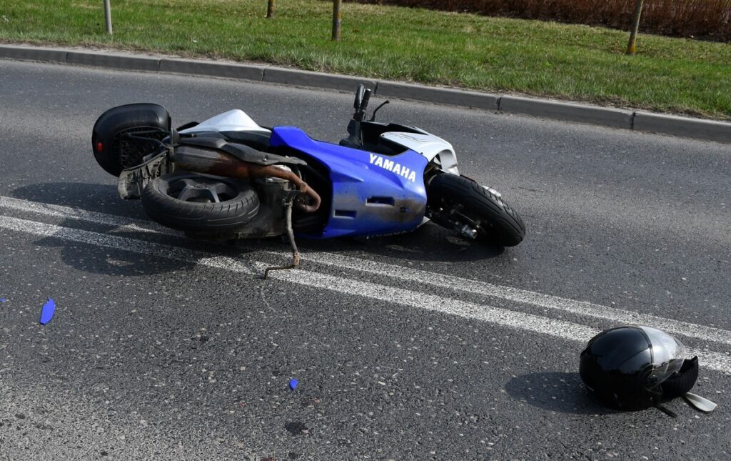 Złożony skuter leży na asfalcie obok czarnego kasku, wzdłuż drogi z wyraźnymi liniami.