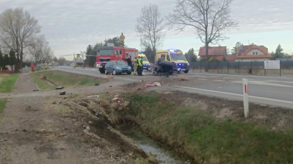 Na drodze leży zniszczony samochód, obok pracują strażacy i ratownicy medyczni, w tle widać śmigłowiec i pojazdy służb.