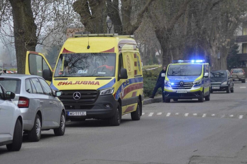 Karetka pogotowia z napisem "AMBULANS" zatrzymana na ulicy, obok policyjny samochód z włączonymi sygnałami świetlnymi.