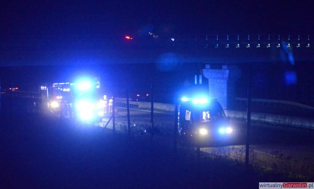 Na drodze w nocy zatrzymane są pojazdy służb ratunkowych z włączonymi niebieskimi światłami.