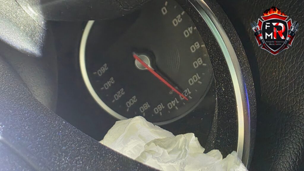 Wnętrze samochodu z widocznym prędkościomierzem, wskazującym 0 km/h, oraz zmiętym papierowym ręcznikiem na desce rozdzielczej.