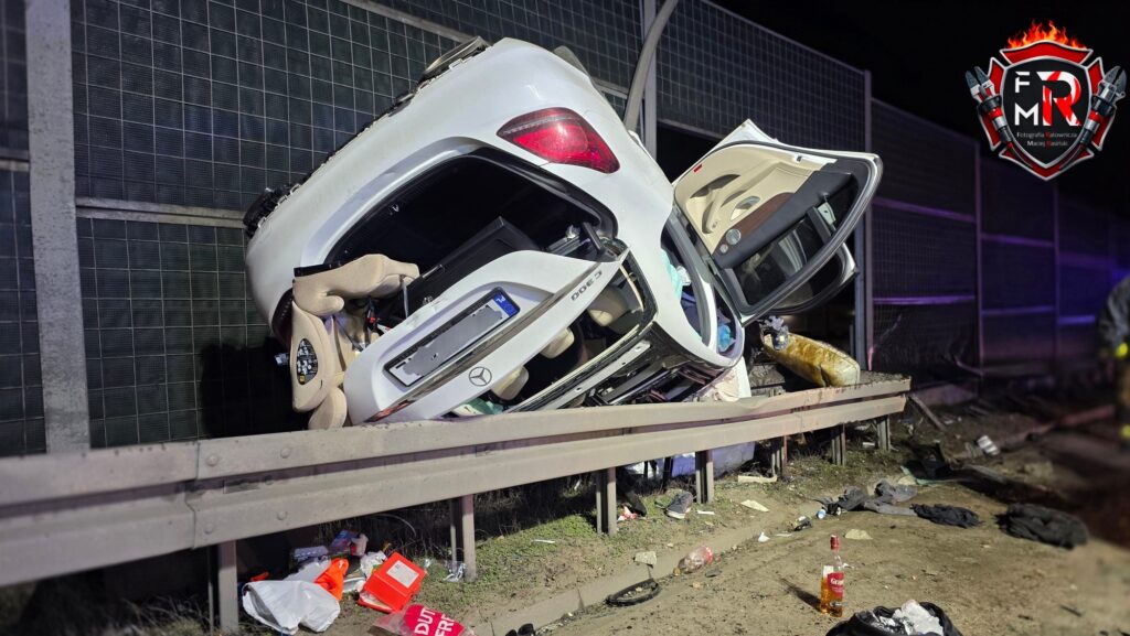 Rozbity biały samochód osobowy leży na boku na barierze ochronnej, wokół porozrzucane śmieci i butelki.