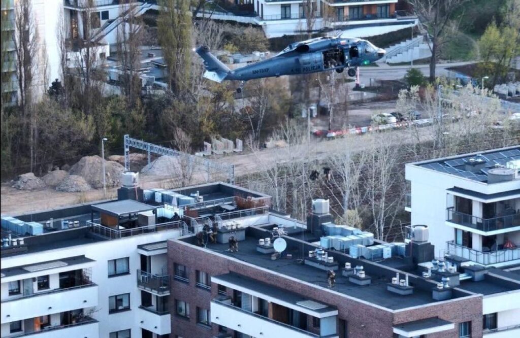 Śmigłowiec policyjny unosi się nad budynkami mieszkalnymi, a na dachach widoczni są funkcjonariusze w mundurach.