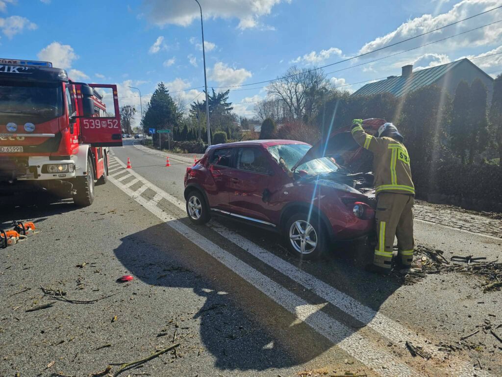 Strażak w mundurze sprawdza uszkodzony samochód osobowy na drodze, obok stoi wóz strażacki.