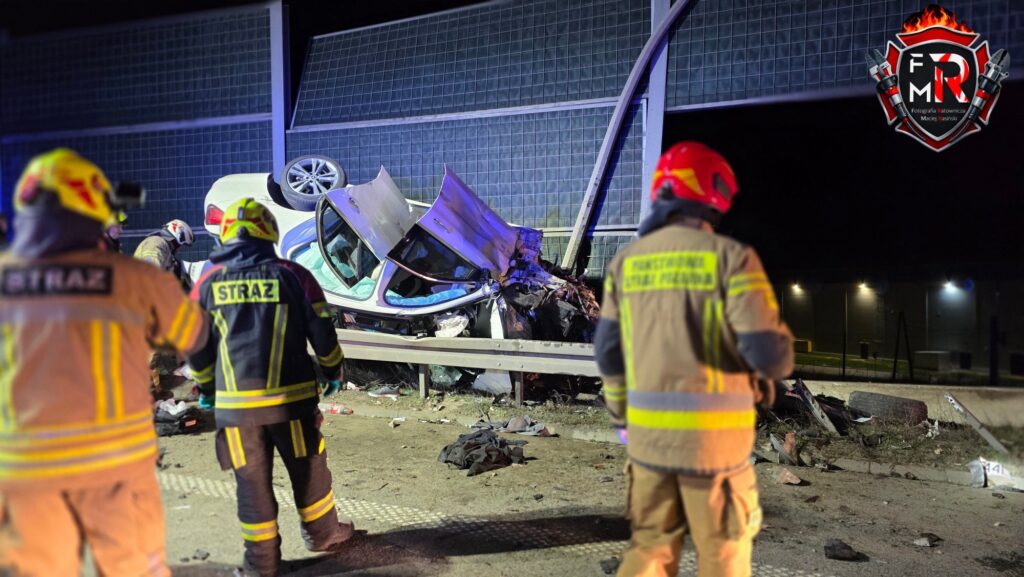 Grupa strażaków ocenia skutki wypadku samochodowego, gdzie pojazd leży na dachu przy barierce.