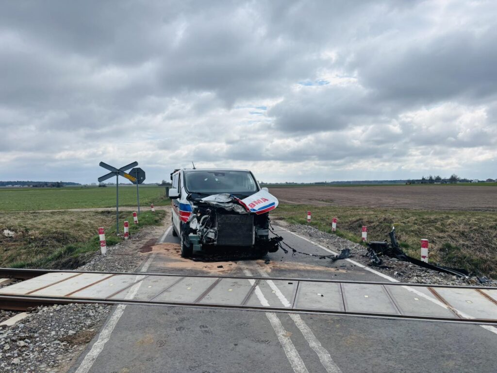 Uszkodzony samochód dostawczy z widocznymi zniszczeniami stoi na przejeździe kolejowym w polu.
