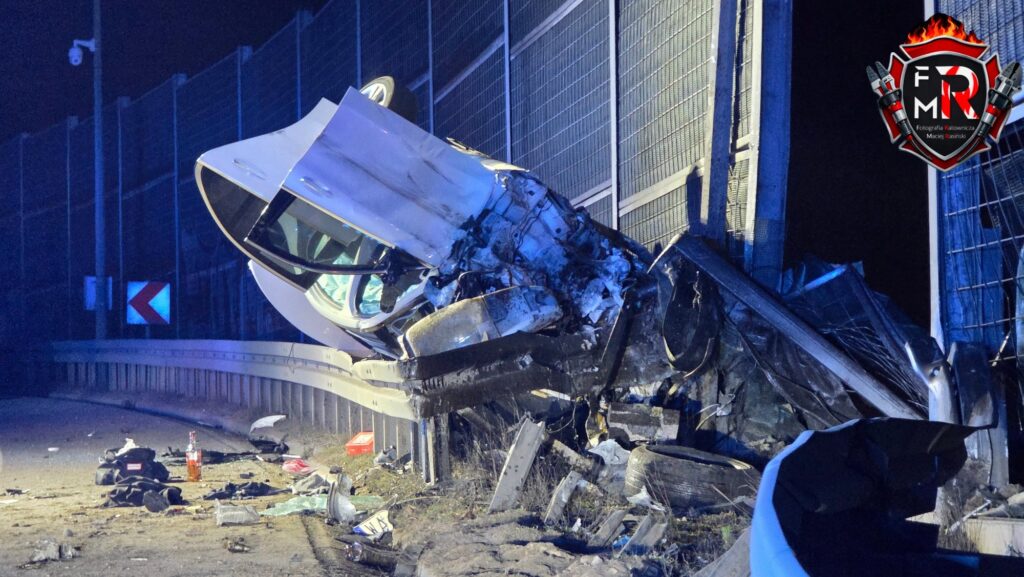 Rozbity samochód osobowy leży na poboczu drogi, oparty o metalowe ogrodzenie, otoczony fragmentami wraku i odpadkami.