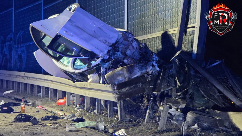 Zniszczony samochód osobowy leży na barierze ochronnej obok drogi, otoczony odpadami i zniszczeniami po wypadku.