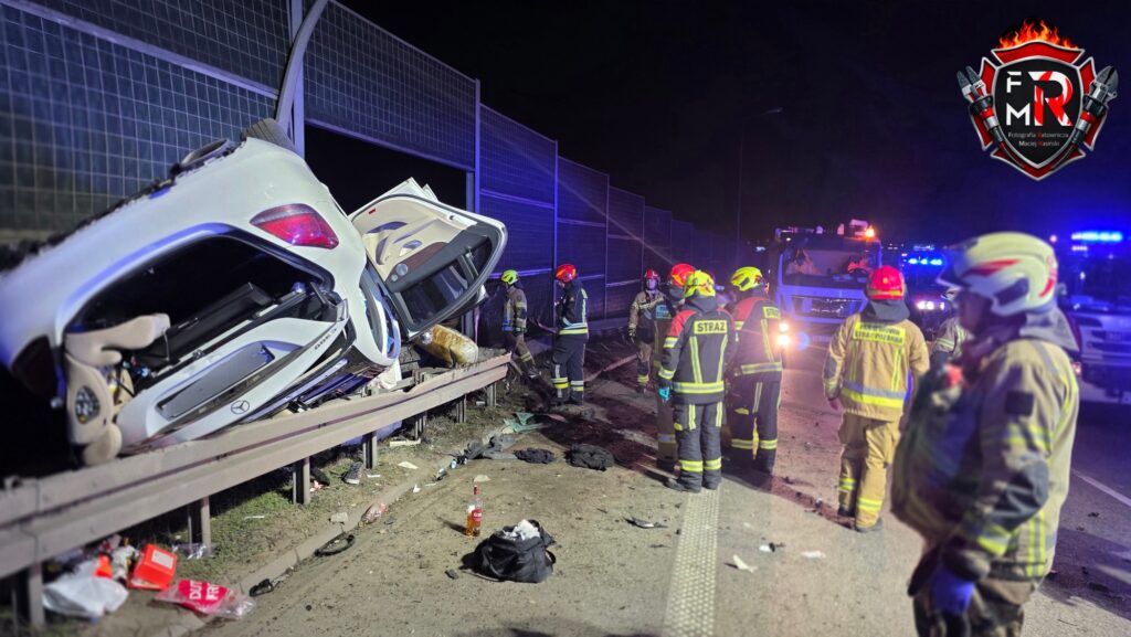 Ratownicy w kaskach i mundurach pracują przy przewróconym samochodzie na poboczu drogi, w nocy.