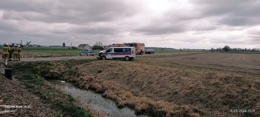 Grupa strażaków w mundurach stoi obok drogi, obok pojazdów ratunkowych i policyjnych, w wiejskim krajobrazie.