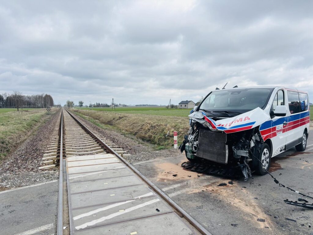 Uszkodzony pojazd dostawczy z widocznym zderzeniem, stojący obok torów kolejowych na otwartym terenie.
