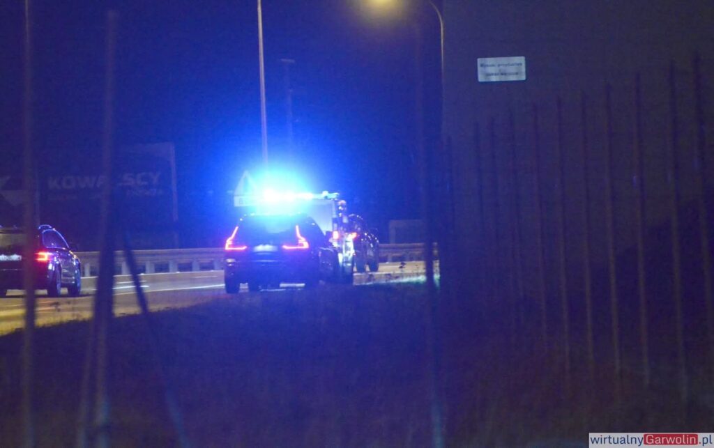 Na drodze w nocy stoi radiowóz z włączonymi sygnałami świetlnymi, obok przejeżdżają inne samochody.