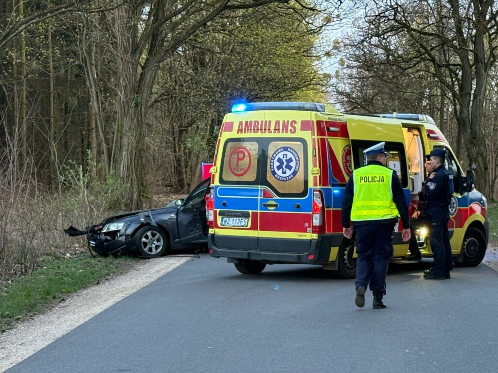 Karetka pogotowia oraz policja na miejscu wypadku drogowego, obok uszkodzonego samochodu w lesistym otoczeniu.