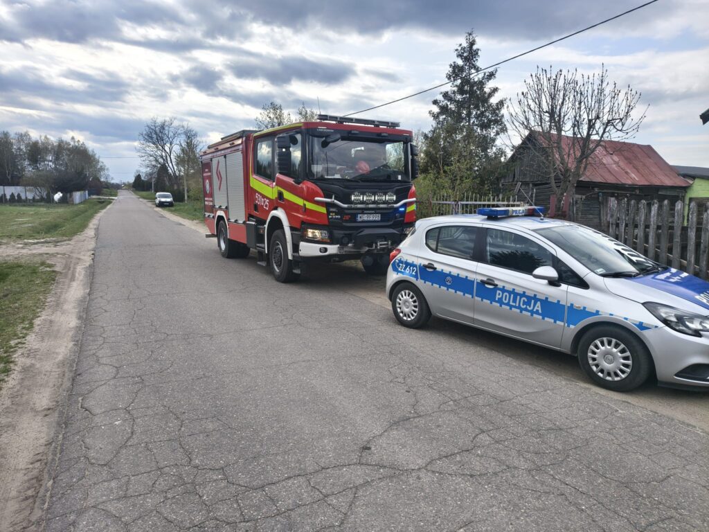 Strażacki wóz bojowy oraz radiowóz policyjny zatrzymane na drodze w wiejskim otoczeniu, obok widocznych domów.