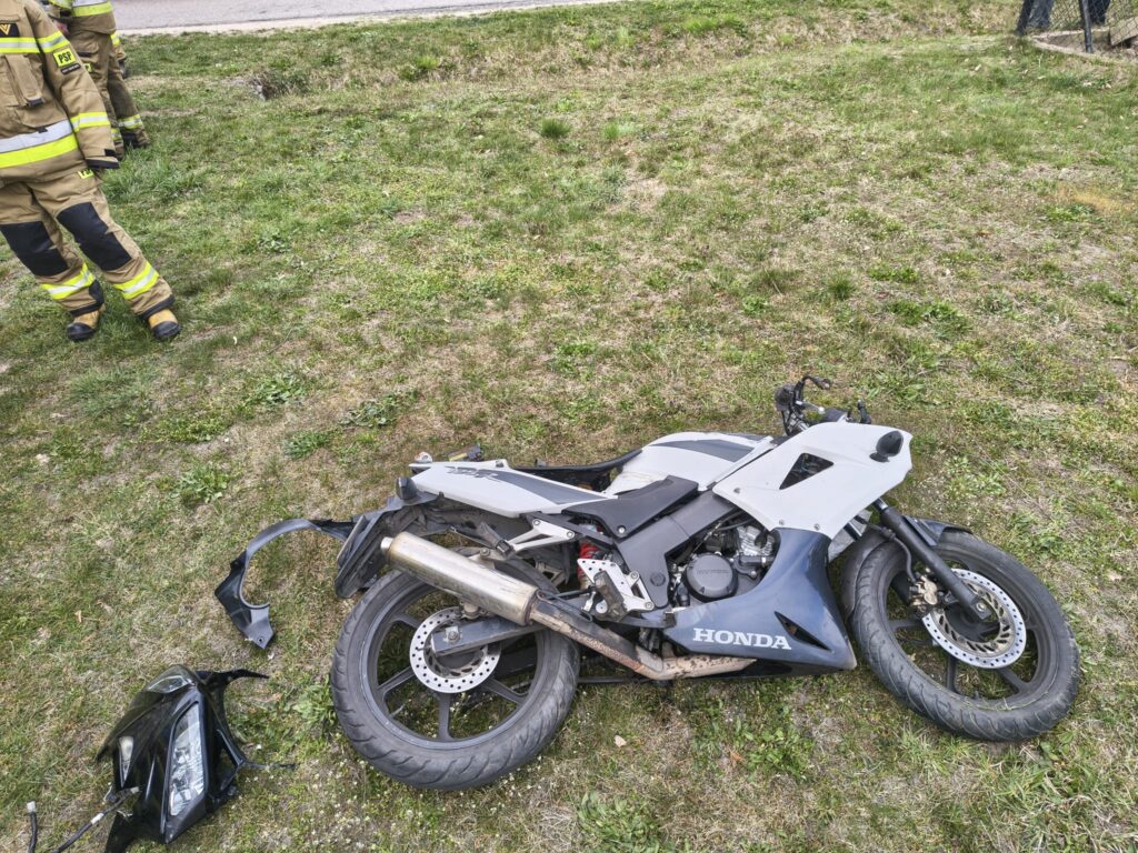 Złomowany motocykl Honda leży na trawie obok drogi, obok niego widoczni strażacy w mundurach.