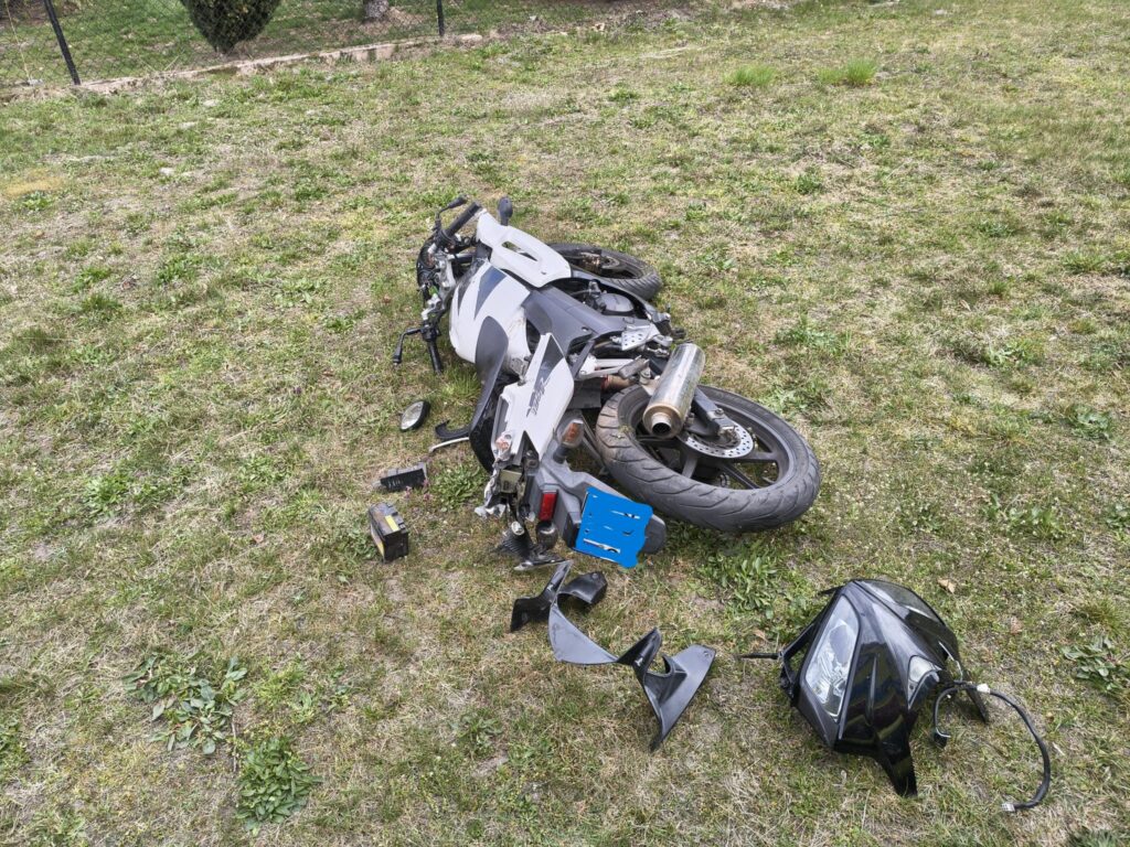 Zniszczony motocykl leży na trawie, obok niego rozrzucone części, w tym lusterka i tylne światło.