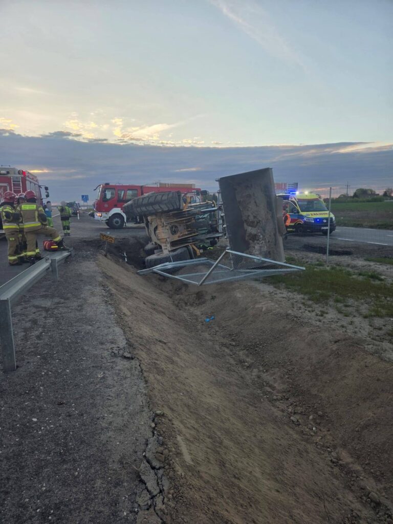 Na drodze leży przewrócony traktor, obok pracują strażacy oraz pojazdy ratunkowe. W tle widać niebo z chmurami.