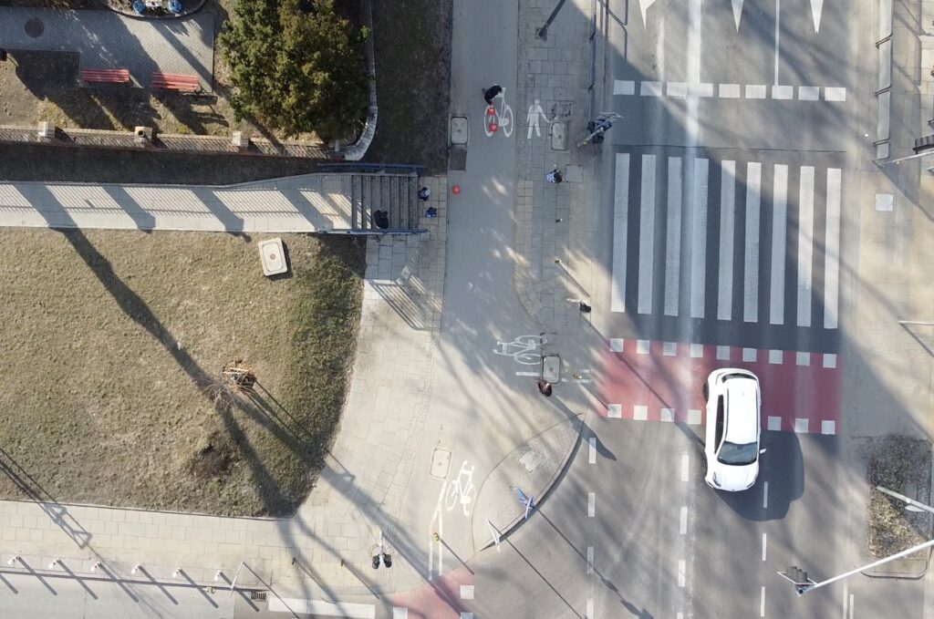 Osoba na rowerze przejeżdża przez ścieżkę rowerową obok przejścia dla pieszych, podczas gdy samochód zbliża się do skrzyżowania.