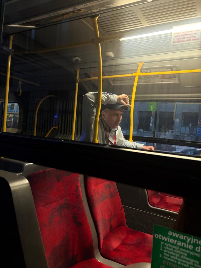 Mężczyzna w czapce stoi w autobusie, opierając rękę na barierce, w pustym wnętrzu z czerwonymi siedzeniami.