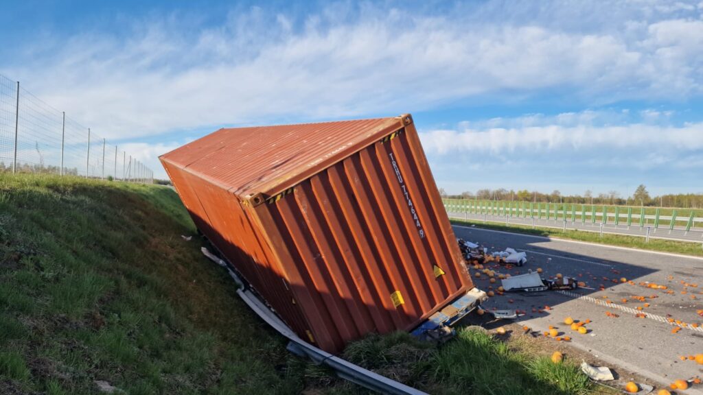 Przewrócony kontener na drodze, obok leżące pomarańcze i resztki ładunku, w tle niebieskie niebo.