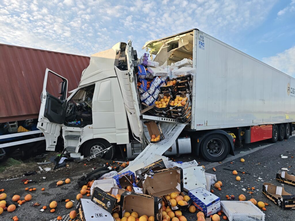 Uszkodzony ciężarowy pojazd z otwartą naczepą, z której wypadły skrzynki z pomarańczami, leżącymi na drodze.