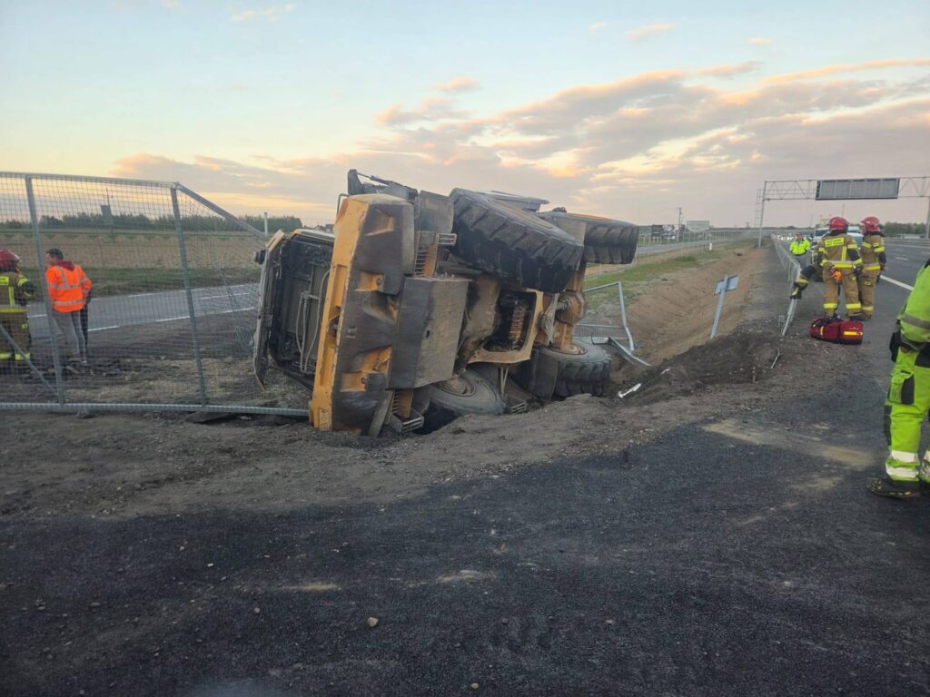 Żółta ciężarówka przewrócona na bok przy drodze, obok grupy strażaków i ratowników medycznych.
