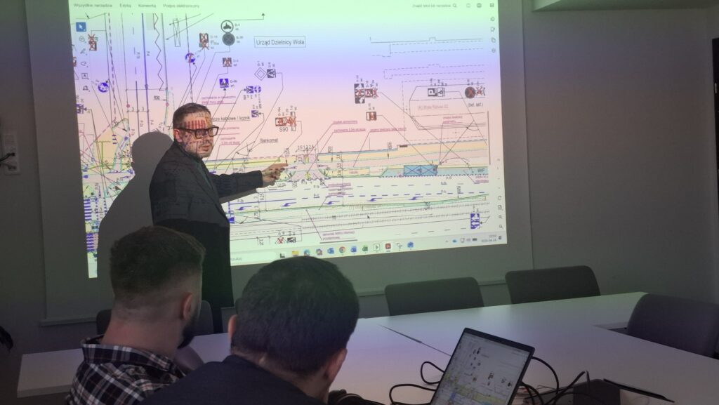 Mężczyzna w okularach wskazuje na projektorze szczegóły schematu, a słuchacze siedzą przy stole w sali konferencyjnej.