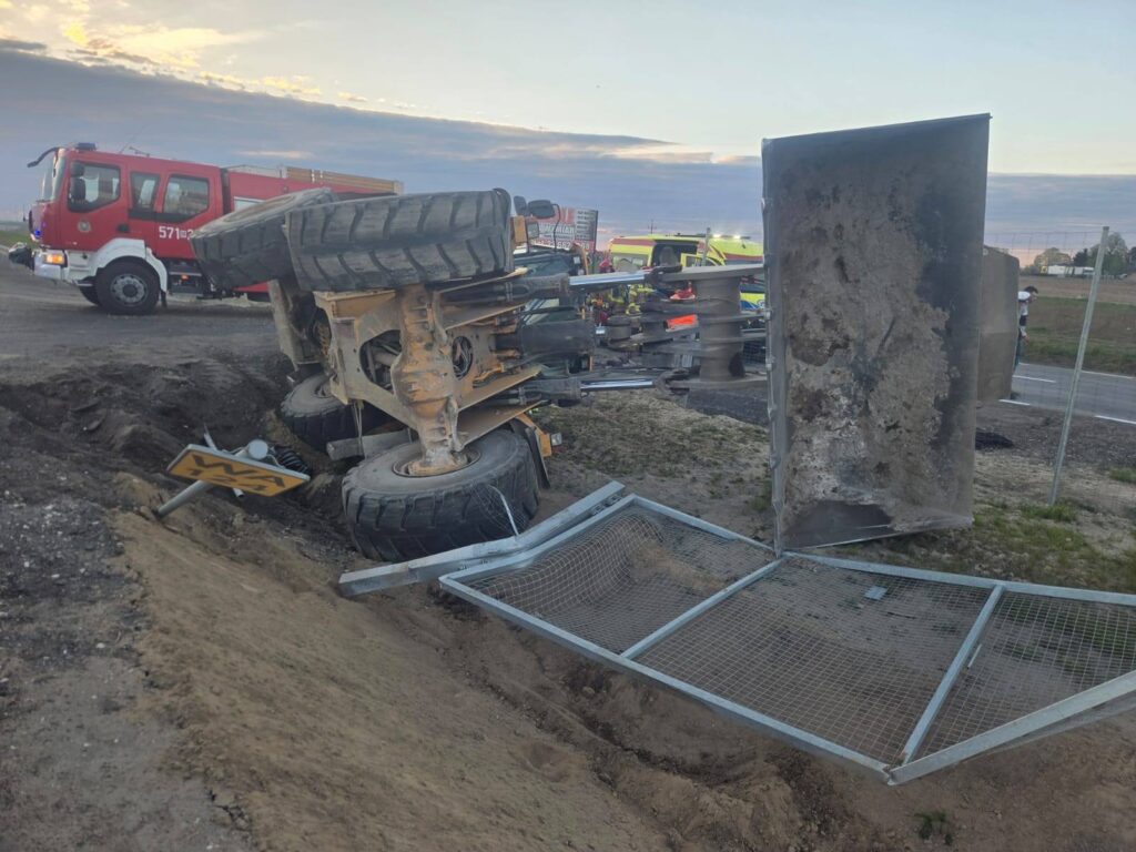 Przewrócony pojazd ciężarowy leży na boku obok drogi, a w tle widoczne są pojazdy służb ratunkowych.