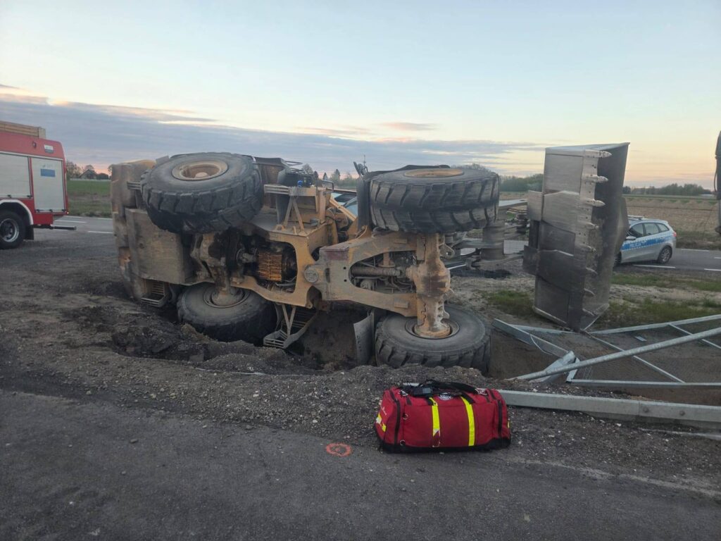 Przewrócony pojazd budowlany z dużymi kołami leży na boku obok drogi, a obok znajduje się czerwona torba ratunkowa.