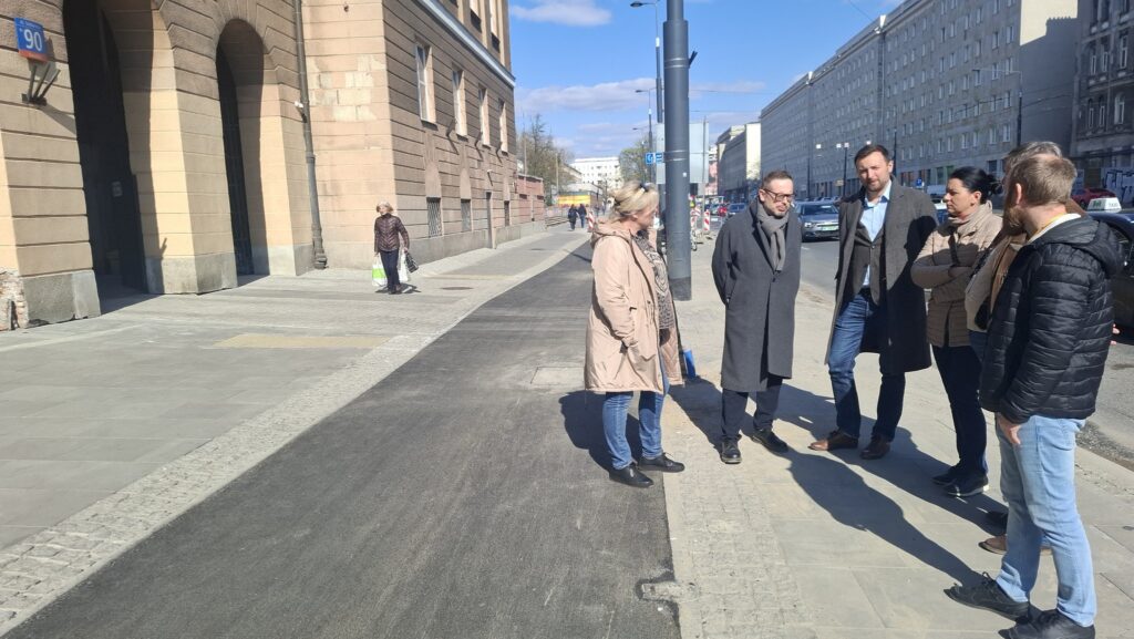 Grupa pięciu osób rozmawia na chodniku obok nowoczesnego budynku, w tle przechodnie i samochody.