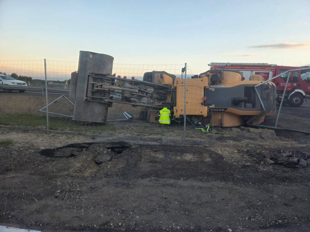 Przewrócony walec drogowy na budowie, obok pracownika w żółtej kamizelce, w tle widoczne pojazdy ratunkowe.