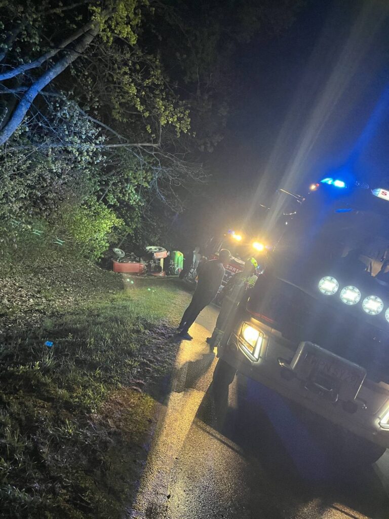 Grupa ratowników pracuje przy przewróconym pojeździe na poboczu drogi w nocy, oświetlona przez latarki i pojazdy służb ratunkowych.