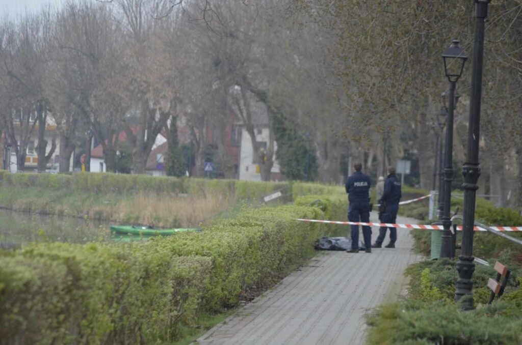 Policjanci zabezpieczają teren przy wodzie, obok zielonego krzewu i chodnika, z widocznym czarnym workiem.