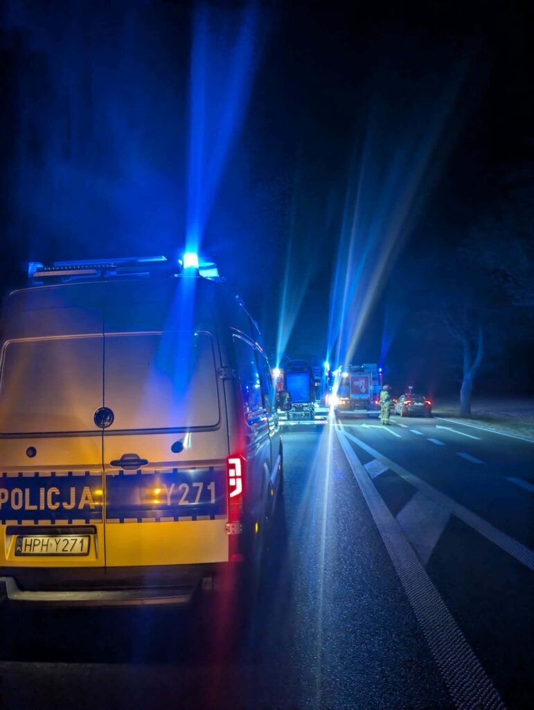 Policyjny radiowóz z włączonymi sygnałami świetlnymi stoi na drodze, w tle widoczne pojazdy służb ratunkowych.