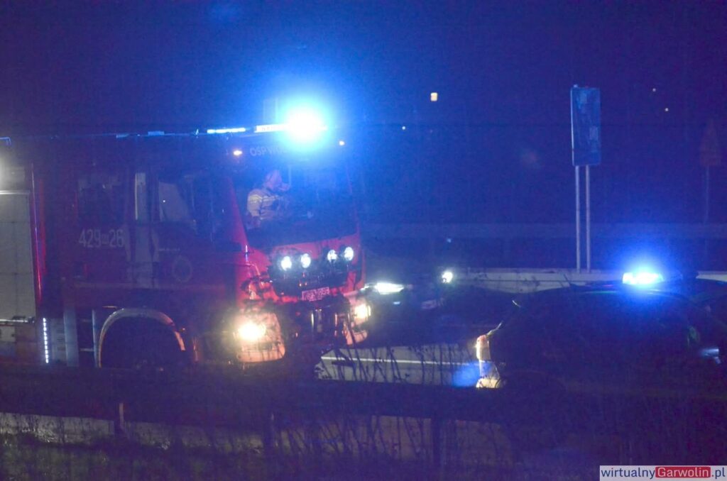 Czerwony wóz strażacki z włączonymi sygnalizatorami świetlnymi zatrzymany przy drodze, obok osobowy samochód.