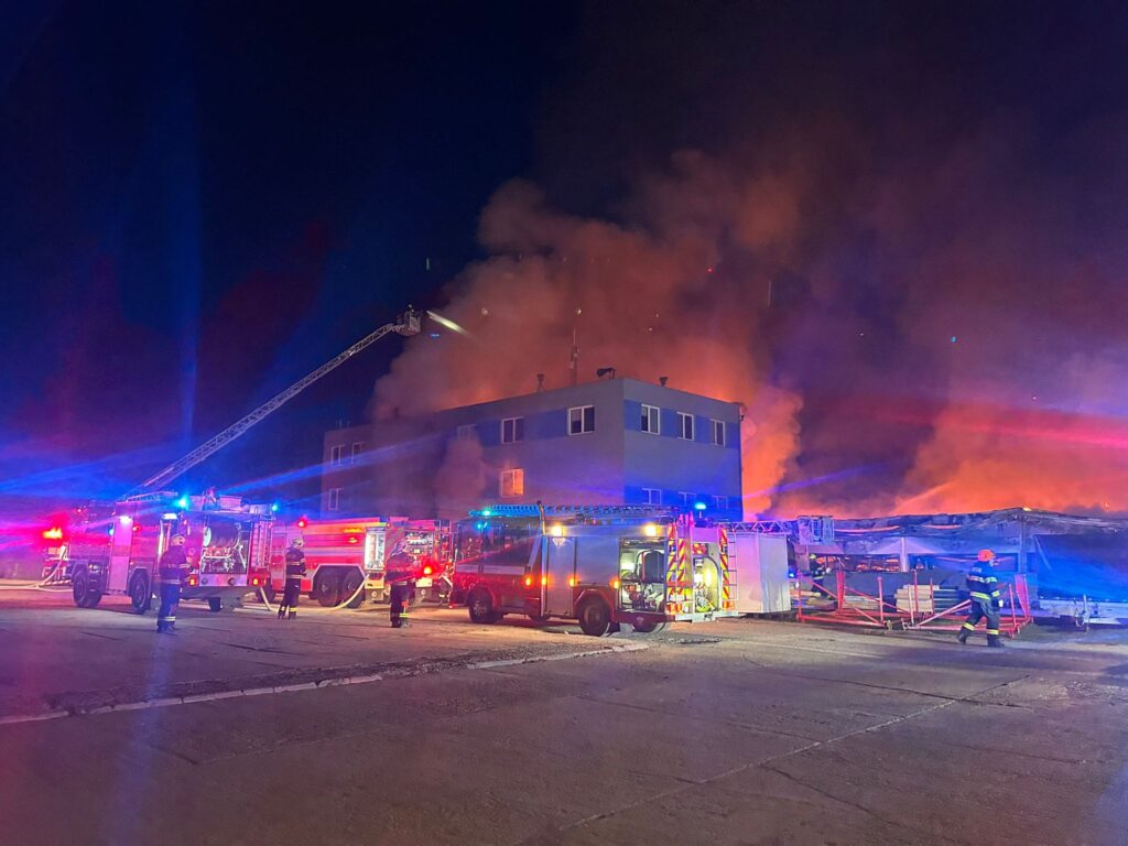 Strażacy z wozami bojowymi gaszą pożar budynku, otoczeni gęstym dymem i jasnymi światłami sygnalizacyjnymi.