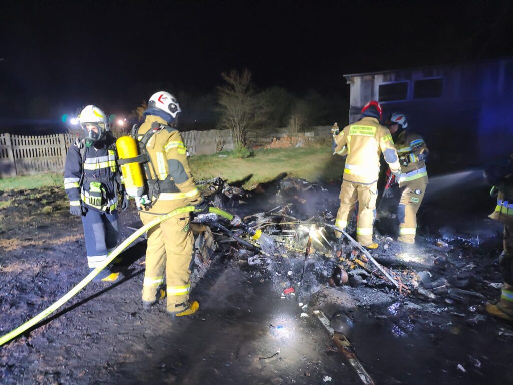 Grupa strażaków w specjalistycznym sprzęcie gaśniczym pracuje nad ugaszeniem pozostałości po pożarze w ciemnej scenerii nocnej.