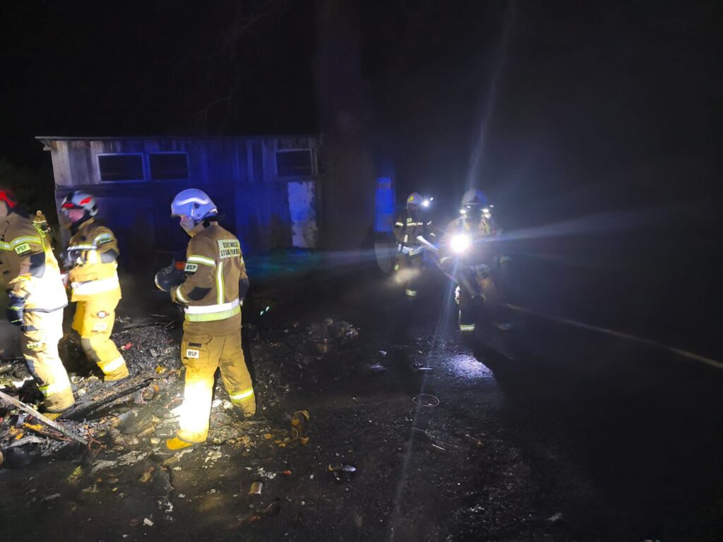 Grupa strażaków w mundurach i hełmach przeszukuje teren po pożarze, oświetlając ciemność latarkami.