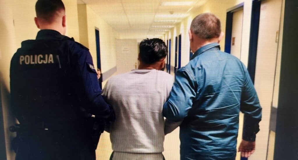 Mężczyzna w szarym dresie jest prowadzony przez dwóch funkcjonariuszy policji korytarzem w budynku.