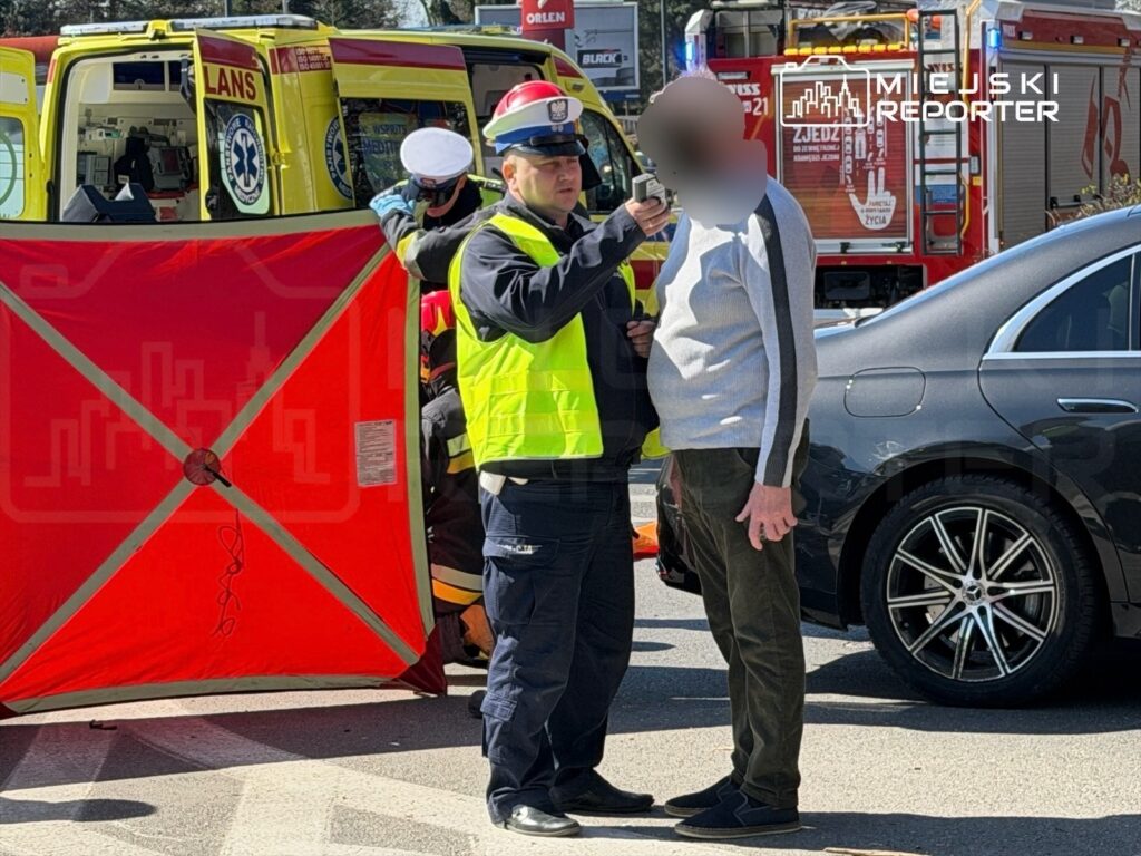 Policjant w kamizelce odblaskowej przeprowadza badanie alkomatem mężczyzny na ulicy, obok czerwonego wozu strażackiego.