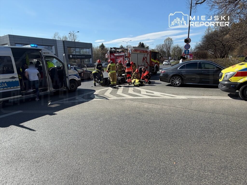 Wielu ratowników medycznych udziela pomocy osobie leżącej na jezdni w pobliżu skrzyżowania, obok pojazdów służb ratunkowych.